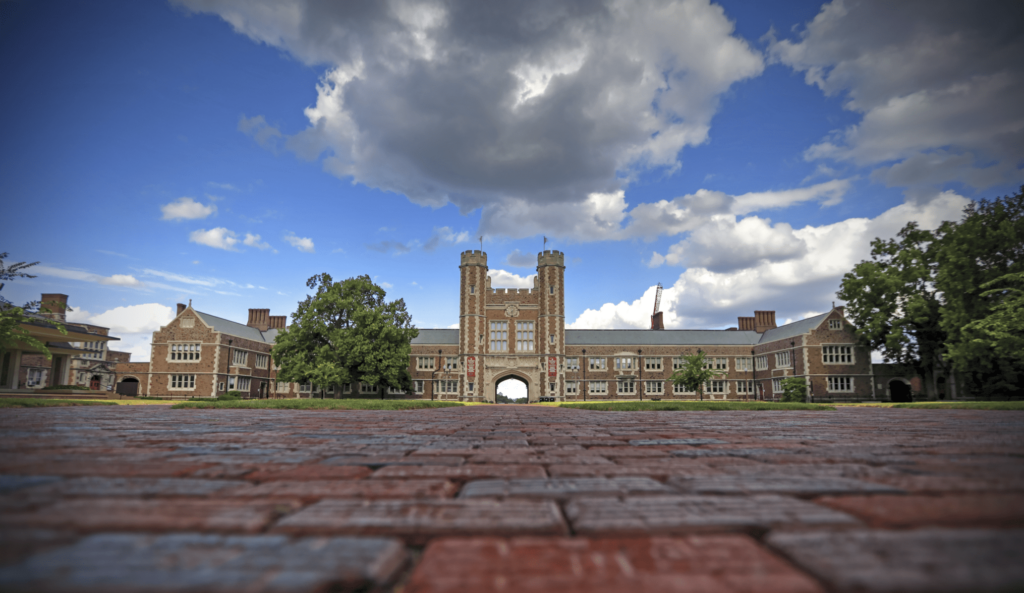 Brookings Hall, a grand red-brick building with a portico and a clock tower. The building is set against a backdrop of partly cloudy blue sky and features stone detailing and arched windows.